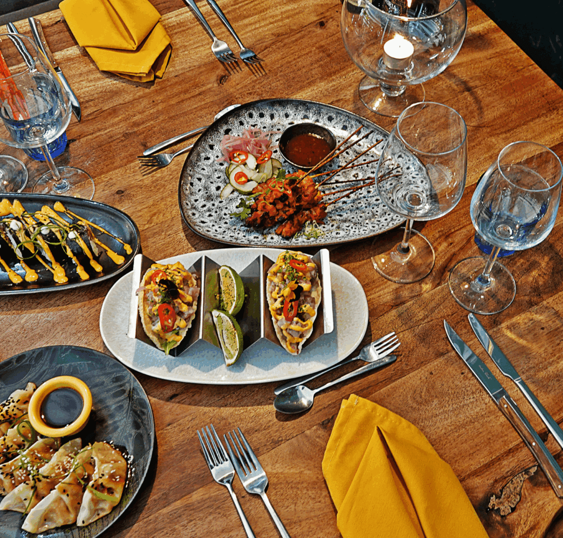 A wooden table set for a meal with plates of assorted appetizers, including tacos, skewers, and dumplings, alongside cutlery, wine glasses, and yellow napkins.